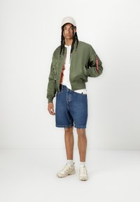 Veste bomber verte, t-shirt blanc, shorts en denim bleu et baskets beiges. Une casquette en acrylique et des cheveux tressés ajoutent des détails à la tenue.