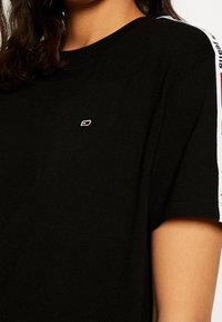 Svart bomullströja med rund hals, korta ärmar, med vita och röda logostripes på axlarna samt en liten broderad logga på bröstet.