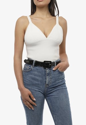 Donna con top bianco senza maniche a coste infilato in jeans blu a vita alta con cintura nera, mano in tasca, su sfondo bianco.