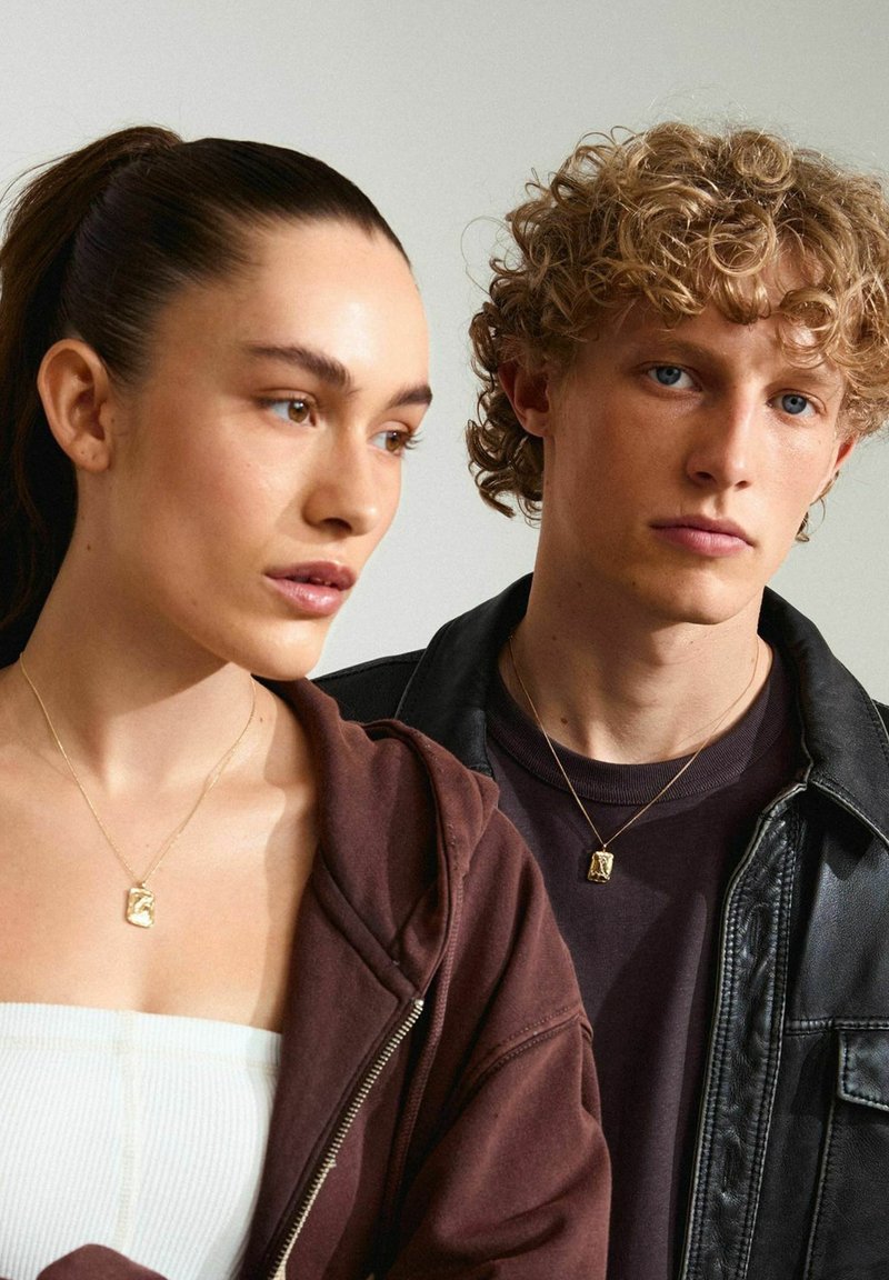 Collar con un colgante de oro; modelo vistiendo una sudadera color burdeos. Chaqueta de cuero texturizada en un modelo masculino; ambos llevan prendas neutras.