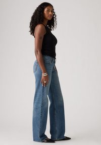 Jeans de mezclilla azul de pierna ancha con un lavado ligeramente desvanecido, cintura alta y diseño de cinco bolsillos. Combinados con una camiseta sin mangas negra.