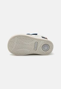Baskets blanches avec une semelle en caoutchouc texturé arborant un motif en zigzag, des accents bleus et des détails de logo sur le talon et la semelle.