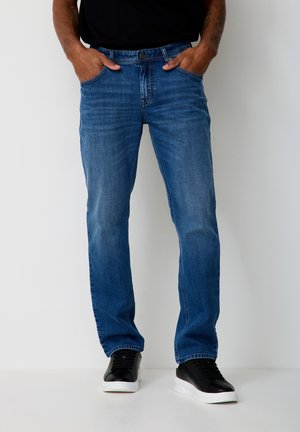 Man wearing blue straight-leg jeans and black sneakers standing with hands in pockets against white background.