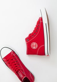 Czerwone płócienne trampki wysokie i niskie z białymi gumowymi podeszwami, kontrastującymi czarnymi noskami i okrągłymi akcentami logo w okolicy kostki.
