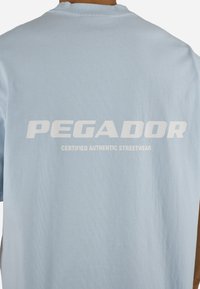 Vaaleansininen T-paita, jonka takana on valkoinen teksti "PEGADOR" ja pienempi teksti "CERTIFIED AUTHENTIC STREETWEAR".