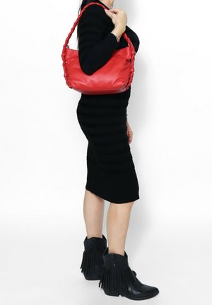 Vrouw in zwarte jurk en zwart franjeslaarzen met een rood leren handtas over haar schouder tegen een witte achtergrond.