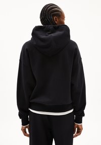 Schwarzer Kapuzenpullover aus weichem Material. Lockerer Schnitt, gerippte Bündchen und Saum. Mit einem weißen Streifen am unteren Rand. Ansicht von hinten.