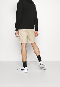 Beiget shortsit, joissa rento leikkaus ja kääritty helm, yhdistetty mustaan huppariin ja valkoisiin Nike-tennareihin. Shortseissa näkyy tekstuuri.