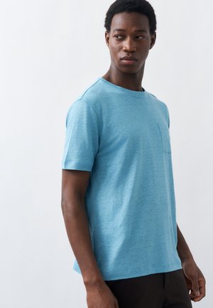 Νέος άνδρας που φοράει ανοιχτό μπλε κοντομάνικο μπλουζάκι με μικρή τσέπη στο στήθος, στέκεται και κοιτάζει προς τα αριστερά του.