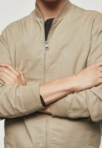 Giubbotto bomber beige chiaro con colletto e polsini a coste, dotato di una cerniera frontale e tessuto strutturato, mostrante braccia incrociate su uno sfondo neutro.