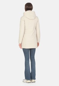 Giacca isolata di un beige chiaro con colletto alto e cappuccio, caratterizzata da un design aderente e maniche lunghe. Indossata con jeans a zampa d'elefante.
