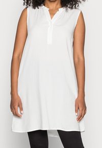 Túnica blanca sin mangas con escote en V, tapeta con botones y dobladillo recto. Hecha de tela ligera, que ofrece una textura suave.