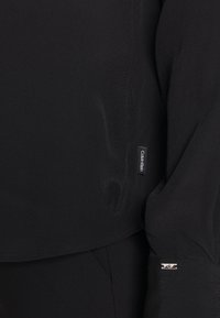 Blusa nera con maniche lunghe, dalla texture liscia e dal fit leggermente rilassato. Presenta un piccolo etichetta Calvinklein sulla manica sinistra.