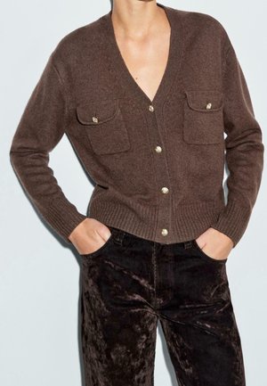 Woman wearing a brown buttoned cardigan with flap pockets and dark crushed velvet pants, hands in pockets, against plain background.