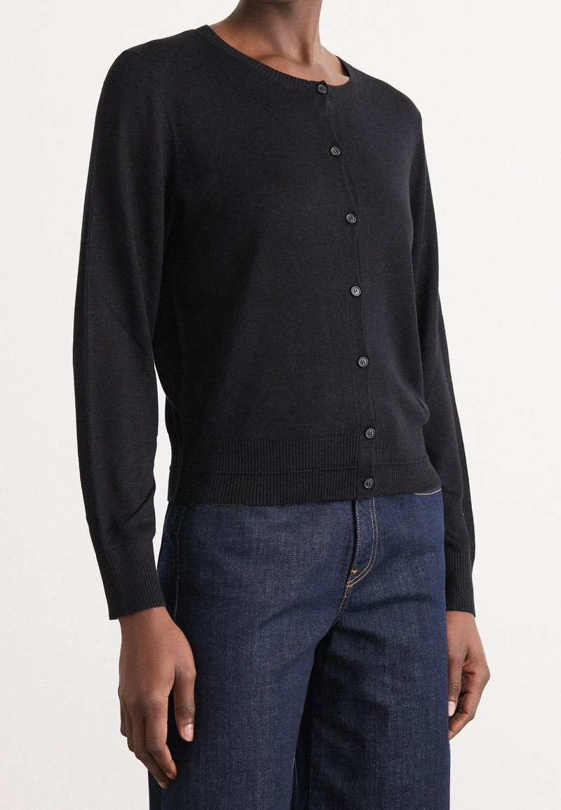 Personne portant un cardigan noir boutonné à manches longues, associé à un jean en denim bleu foncé, sur un fond clair uni.