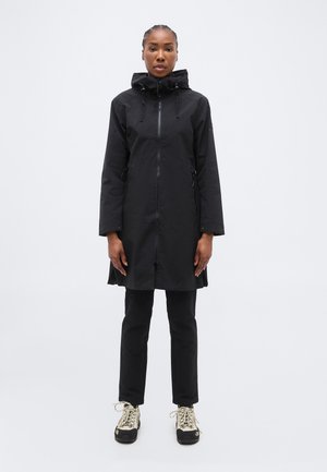 Nő fekete térdig érő kapucnis kabátban, fekete nadrágban és bézs túracipőben, szemben áll egy egyszínű háttér előtt.