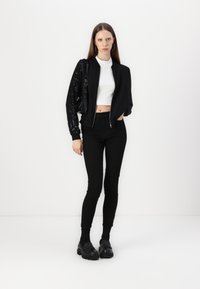 Schwarze Bomberjacke mit Paillettenärmeln, weißer kurzer Rollkragenpullover, schwarze Slim-Fit-Hosen und schwarze Schuhe. Schlichte Tasche mit Hardware-Akzenten.