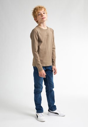 Beige Langarm-T-Shirt mit kleinem Logo, kombiniert mit blauen Jeans und weißen Sneakern mit schwarzen Akzenten. Glatter Stoff und lässige Passform.
