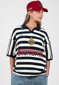 Femme portant un polo rayé bleu marine et blanc avec le logo "Prohibited", un emblème étoile et une casquette rouge avec texte et logo brodés.