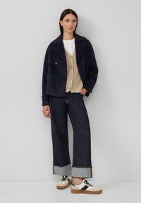 Vrouw in navy pea coat, beige gebreide vest met knopen over een witte blouse, brede donkere jeans met omslag en witte sneakers met zwarte strepen.