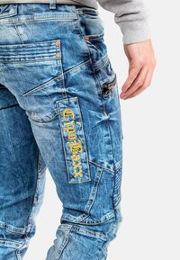 Jeans in denim di un blu chiaro con una finitura sbiadita, caratterizzati da più tasche, cuciture a contrasto e un logo ricamato sul lato.