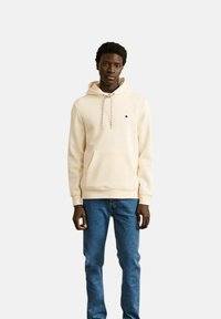 Joven con una sudadera color crema con cordones y jeans azules, de pie contra un fondo liso claro.