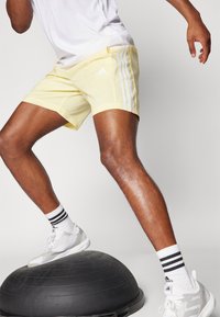 Ανοιχτό κίτρινα αθλητικά σορτς με λευκή τριπλή λωρίδα, συνδυασμένα με λευκά αθλητικά παπούτσια και κάλτσες με μαύρες λωρίδες, πάνω σε εργαλείο ισορροπίας.