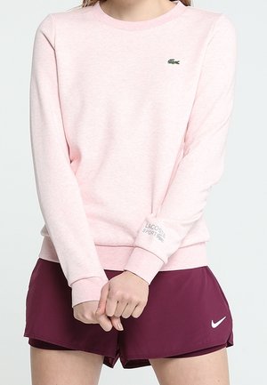 Persoon die een lichtroze Lacoste Sport sweatshirt en donkerpaarse Nike sportshorts draagt, staand met handen gevouwen voor zich.