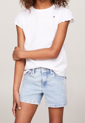 Ung person iført en hvid t-shirt med flæser på ærmerne og lyseblå denimshorts, stående med den ene arm krydset over den anden.