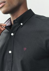 Camisa negra de botones con cuello y botones blancos. Presenta un logo de corona bordado en rosa y detalles de cuadros en el interior del cuello.