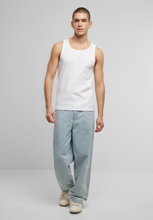 Weißes Baumwoll-Top ohne Ärmel mit rundem Ausschnitt, kombiniert mit hellblauen, weiten Jeans. Die Schuhe sind beiges und weißes Sneakers. Neutraler Hintergrund.