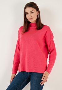 Pull en rose avec un col montant, des manches longues et des fentes sur les côtés. Possède une texture tricot lisse et une coupe décontractée, associé à un jean foncé.