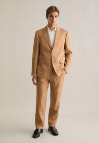 Helles braunes Leinenanzug mit einem einstärkigen Sakko, Revers, flachen Hosen und einem weißen Hemd darunter. Schwarze Schuhe.
