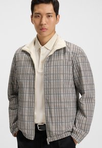 Jachetă ușoară cu un model houndstooth în negru și bej, având un guler crem, închidere cu fermoar și manșete elastice.