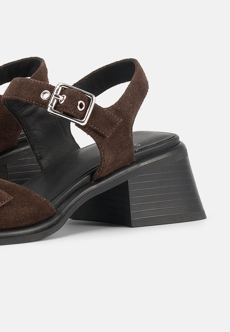 Braune Wildleder-Riemchensandale mit verstellbarem silbernem Schnallenriemen und schwarzem Blockabsatz vor weißem Hintergrund.