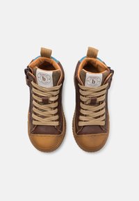 Hnědé kožené tenisky pro batolata s hnědými gumovými podrážkami, béžovými tkaničkami, bočními zipy a značkovými štítky na jazyku, vystavené z pohledu shora na bílém pozadí.