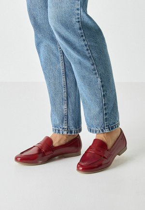 Red leather loafers with smooth texture, featuring a strap across the vamp and low-profile sole, paired with blue jeans.