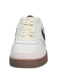 Zapatilla blanca de cuero con logo negro, detalles de gamuza beige y cordones redondeados. Presenta perforaciones en la puntera. Suela de goma.