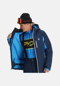 Marineblaue Skijacke mit einer strukturierten Außenseite, Frontreißverschluss und Kapuze. Das Innere weist ein leuchtend blaues Futter und mehrere Taschen auf.