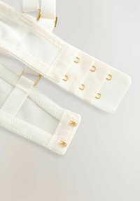 Des sangles en tissu couleur crème avec des sections en mesh et des accessoires en métal doré. Comprend plusieurs fermetures à crochets et à œil pour un ajustement réglable.