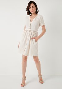 Beige short-sleeve dress features a V-neck, front buttons, and a gathered waist with pockets, made of lightweight fabric.
