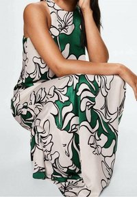 Femme assise avec le coude sur le genou, portant une robe sans manches à grande imprimé floral blanc et vert.