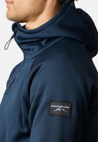 Felpa con cappuccio blu navy, colletto alto, cordino regolabile e una toppa nera cucita con la scritta "SWEDEMOUNT" e grafiche di montagne.