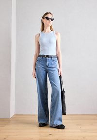 Lys blå ribbet tanktop kombinert med vide denimjeans. Modellen holder en svart veske og har på seg solbriller. Glatte teksturer og avslappet passform.