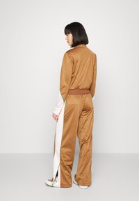 Woman standing facing away, wearing a brown tracksuit with pink and white side stripes and white sneakers against a plain background.