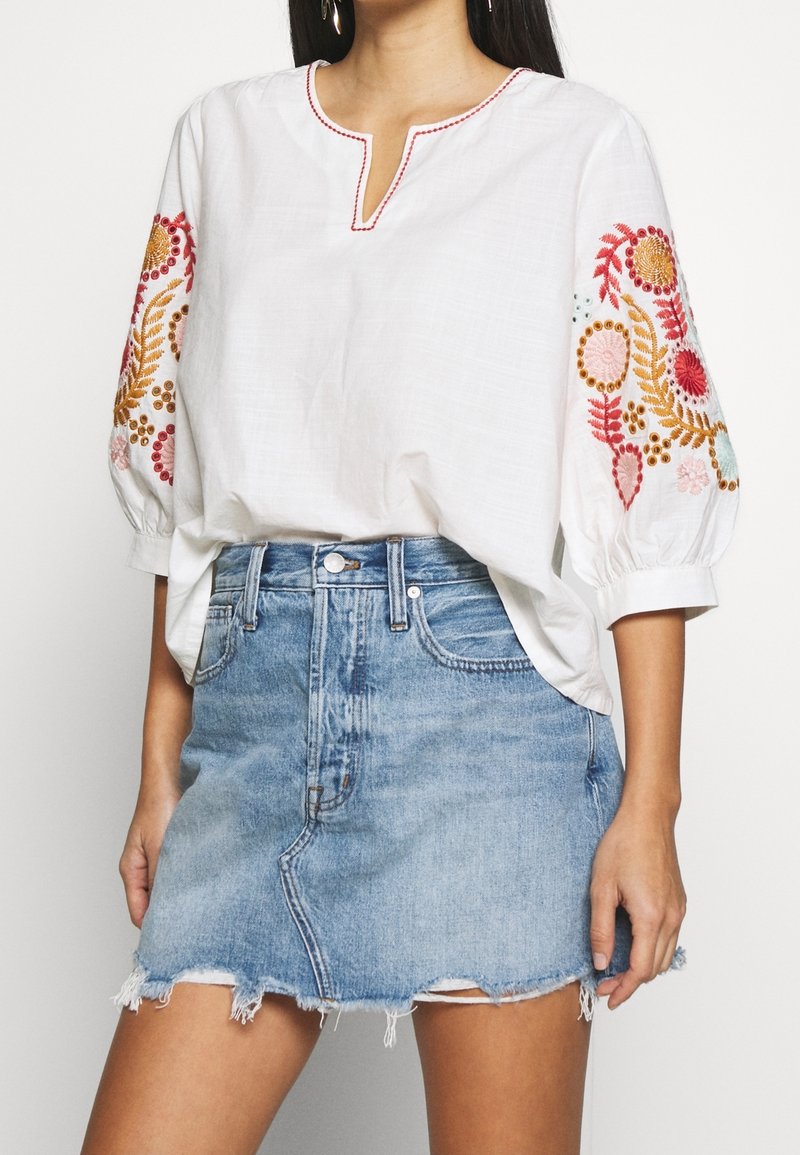 Top in lino bianco con ricami floreali colorati sulle maniche, abbinato a una gonna in denim azzurro chiaro con orlo sfilacciato e tasche frontali.