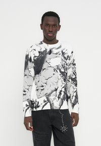 Πλεκτό πουλόβερ σε λευκό με εκφραστικό μαύρο και γκρι σχέδιο. Λαιμόκοψη γύρω, μακριά μανίκια, μανσέτες με ριμπ υφή, φορεμένο με μαύρο τζιν με σκισίματα.