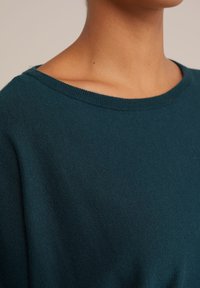 Turkizna pulover z širokim čolnastim izrezom in rebrastim ovratnikom. Mehka tekstura s ravnim krojem, ki prikazuje bližnji posnetek tkanine.