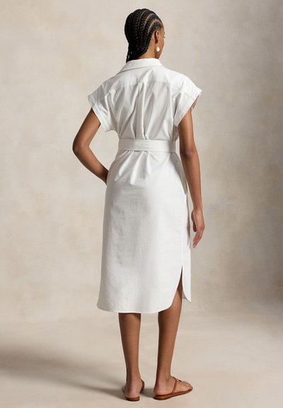 Woman with braided hair wearing a belted white short-sleeve midi dress and brown flat sandals, standing against a plain beige background.