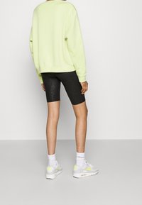 Lichtgroene oversized sweatshirt, zwarte gebloemde biker shorts en witte sneakers met gele accenten, gezien vanaf de achterkant tegen een neutrale achtergrond.